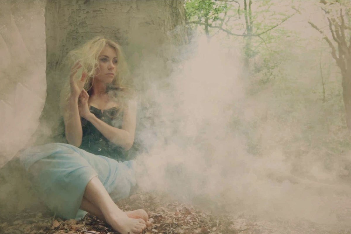 Woman in a blue dress sitting against a tree trunk in a misty, smoke-filled forest, creating an atmospheric fog effect