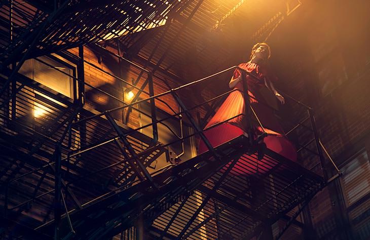 Film noir-style photo of a man in a trench coat and fedora on a fire escape, backlit in warm light