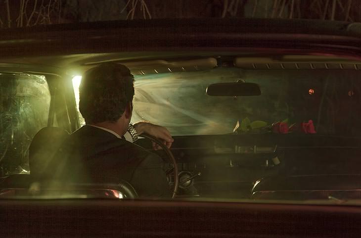 Film noir-style photo of a man in a fedora sitting in a car at night, backlit through the windshield