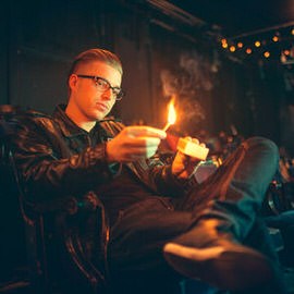 A man in glasses lighting a match in a warmly lit, low-light environment, behind-the-scenes photo from an environmental portrait shoot of a musician