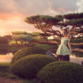 A woman in a green dress standing near a twisted tree beside a pond at a colorful sunset, illustrating how to enhance a beautiful sunset in Photoshop