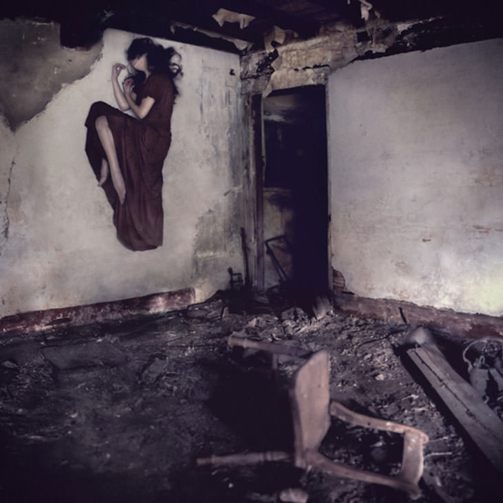 Woman in a long dark red dress floats sideways against the wall of a derelict room with a broken chair on the floor