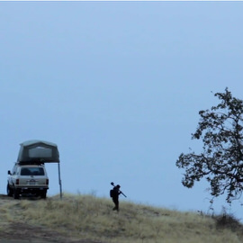 Silhouette of a photographer on a hillside beside a rooftop tent and vehicle at dusk, illustrating travel photography gear suggestions