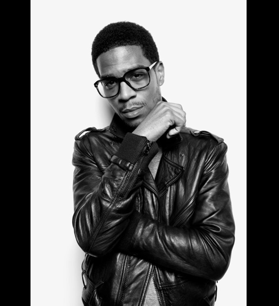 Black-and-white portrait of Kid Cudi in thick glasses and a leather jacket, hand at his chin, on white