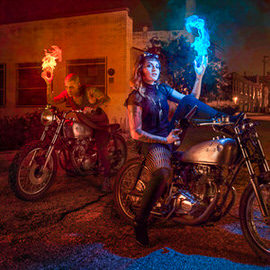 Composite of a blue-skinned figure with glowing fire in each hand standing beside motorcycles at night, demonstrating a realistic photo composite with visual effects