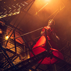 Woman in a red dress dramatically posed on a fire escape with moody golden backlighting, example for creating film noir coloring and lighting in Photoshop