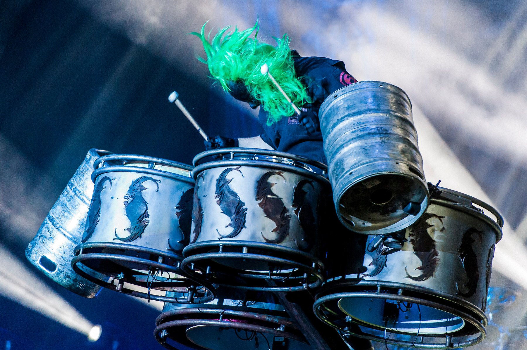 Drummer with bright green hair plays a kit of metal keg drums on a stage under white lights
