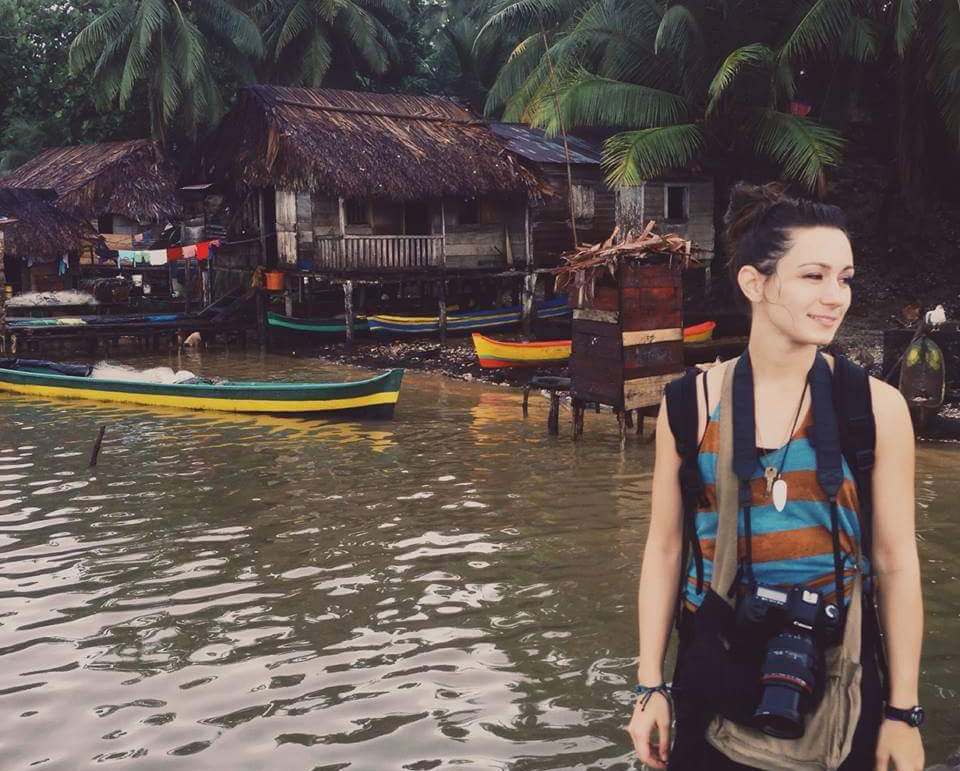Woman with a camera on her shoulder strap stands by the water in front of thatched stilt houses and painted boats
