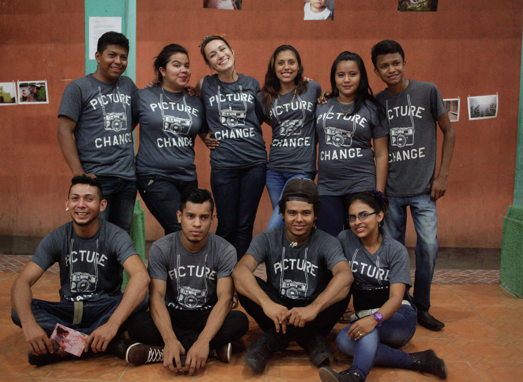Ten young people in matching grey Picture Change T-shirts pose in two rows against an orange wall hung with photos