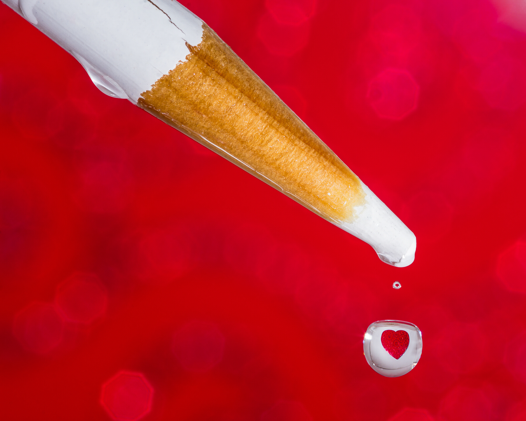Macro of a water drop falling from a white colored pencil against red, a red heart refracted inside it