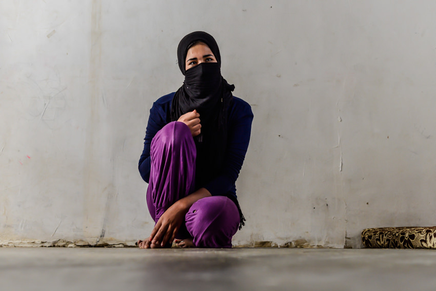 Woman in a black headscarf with her face covered crouches against a bare white wall