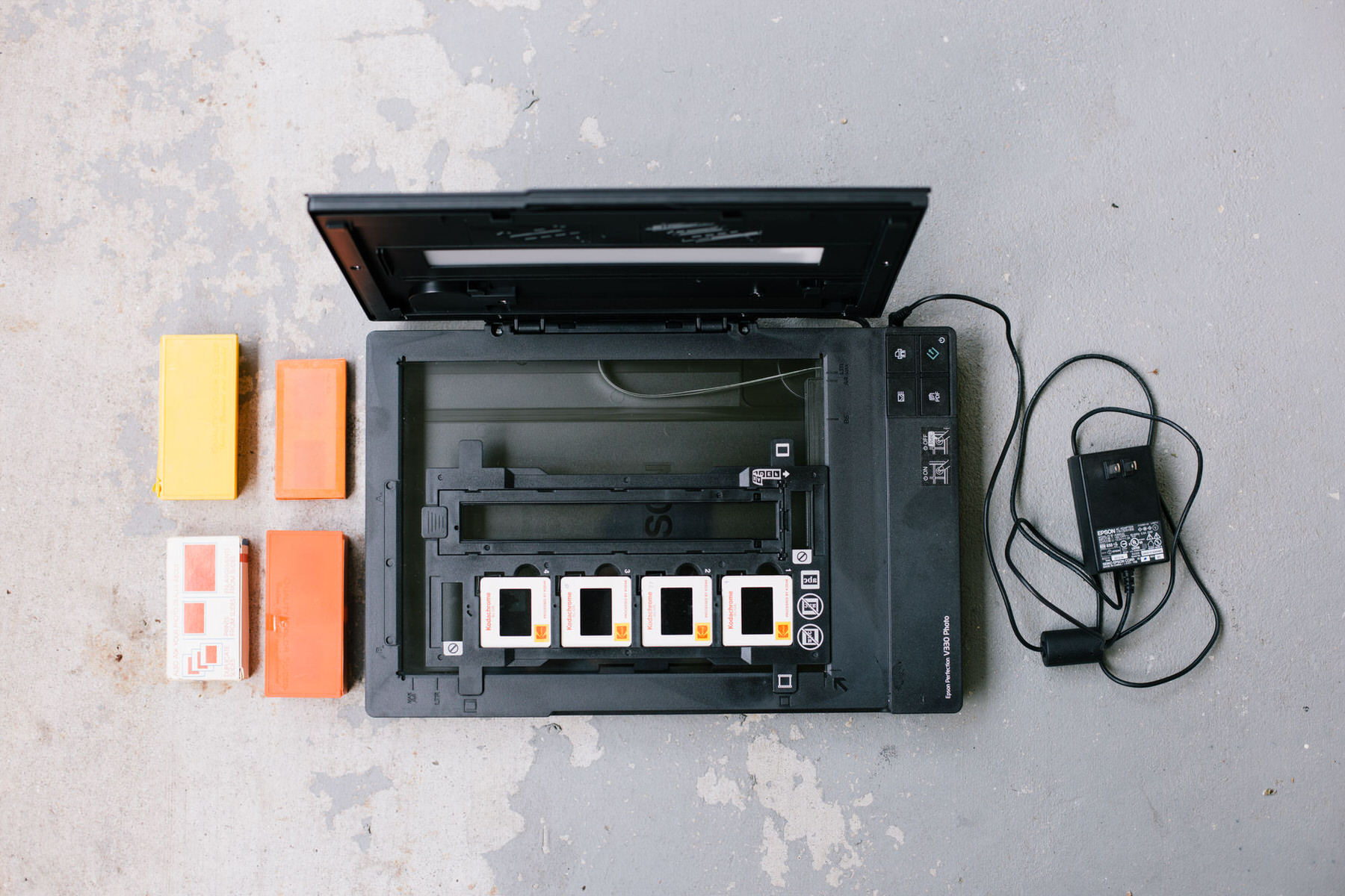 Open flatbed scanner with four slides in its holder, next to slide boxes and a power adapter, seen from above