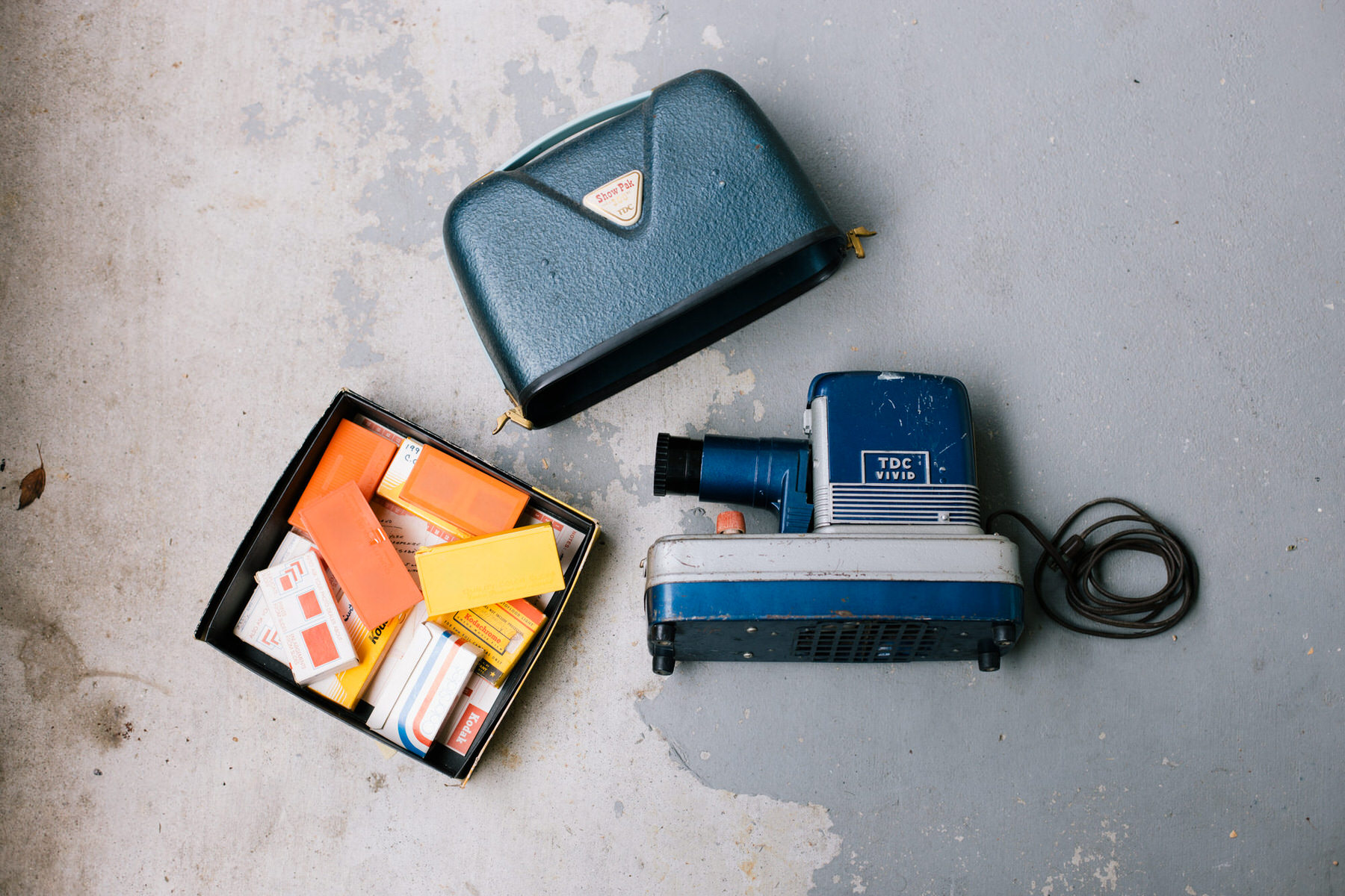 Vintage blue TDC Vivid slide projector beside its blue carrying case and a box of orange and yellow slide cartons