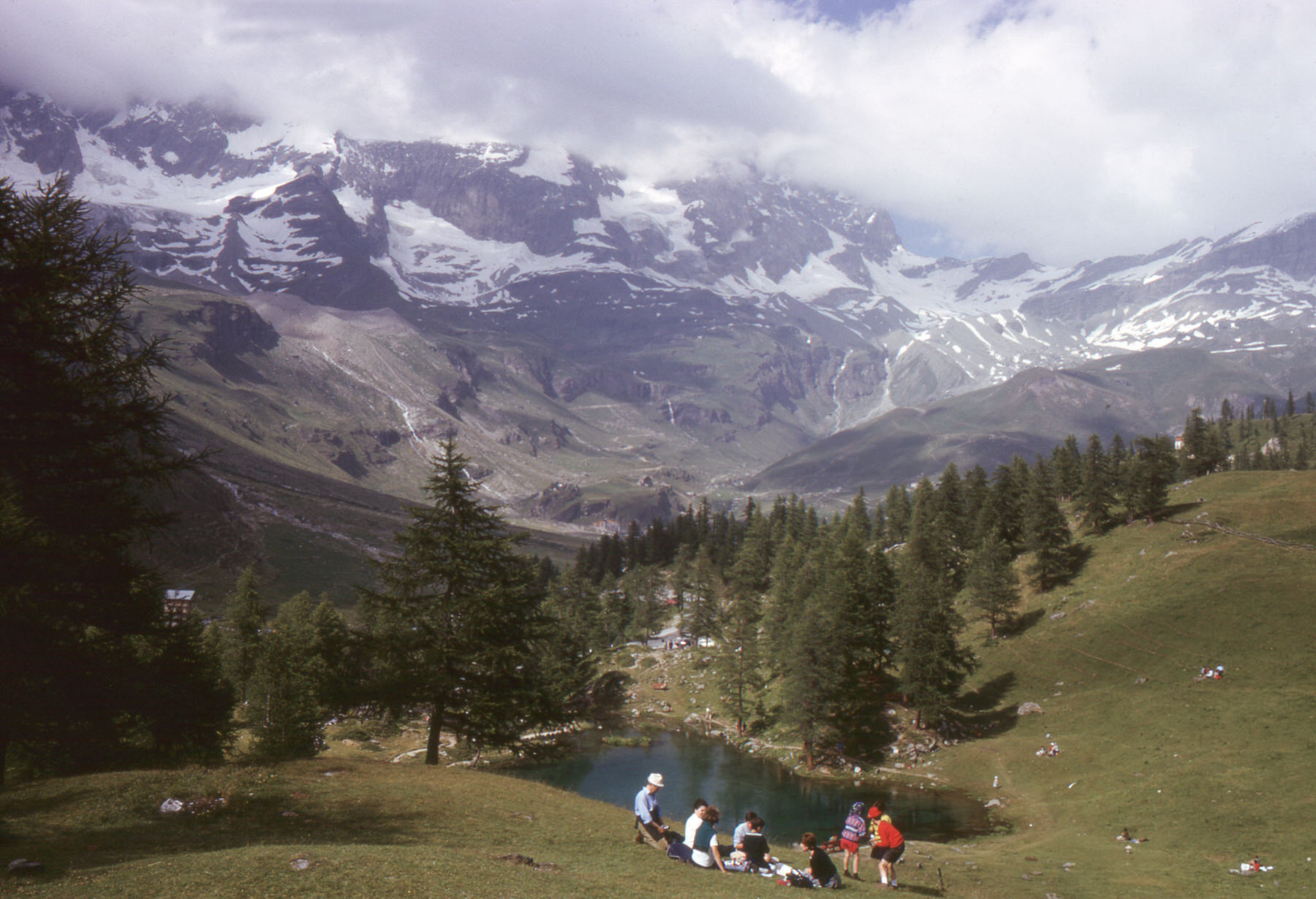 Old color slide of a group picnicking by a small lake among pines below snowy, cloud-capped mountains