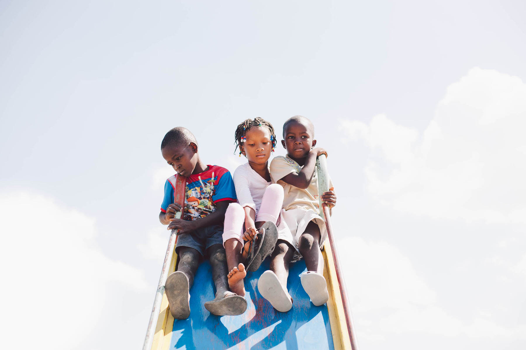 Justin Eisner's photo of three children in Haiti sitting together at the top of a blue playground slide against a pale sky