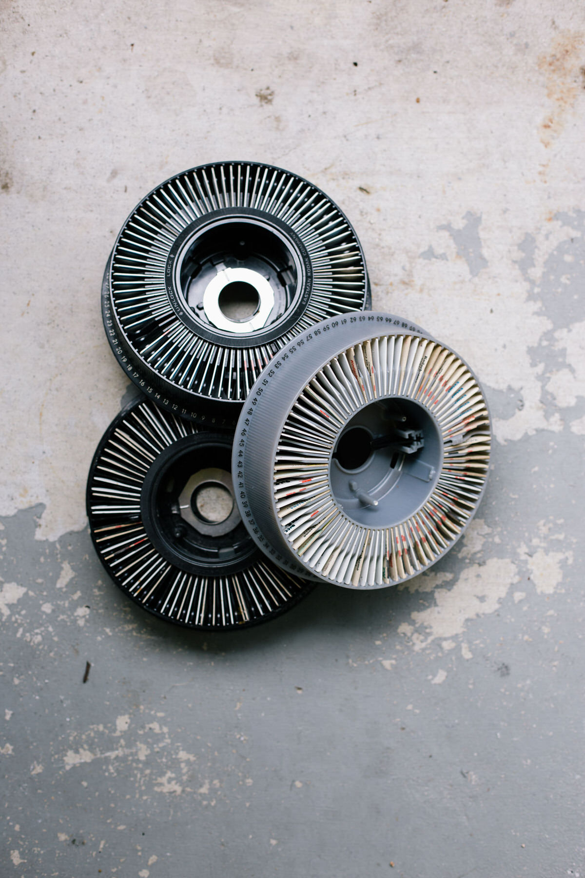 Three round slide carousel trays, two black and one gray, stacked on a worn concrete floor