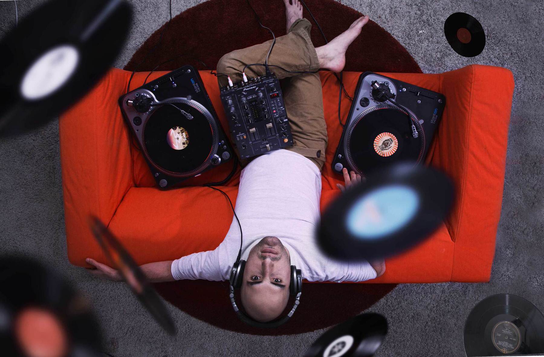 Keean Afrasiabi self-portrait: a man in headphones lying on an orange couch between two turntables and a mixer, vinyl records flying around