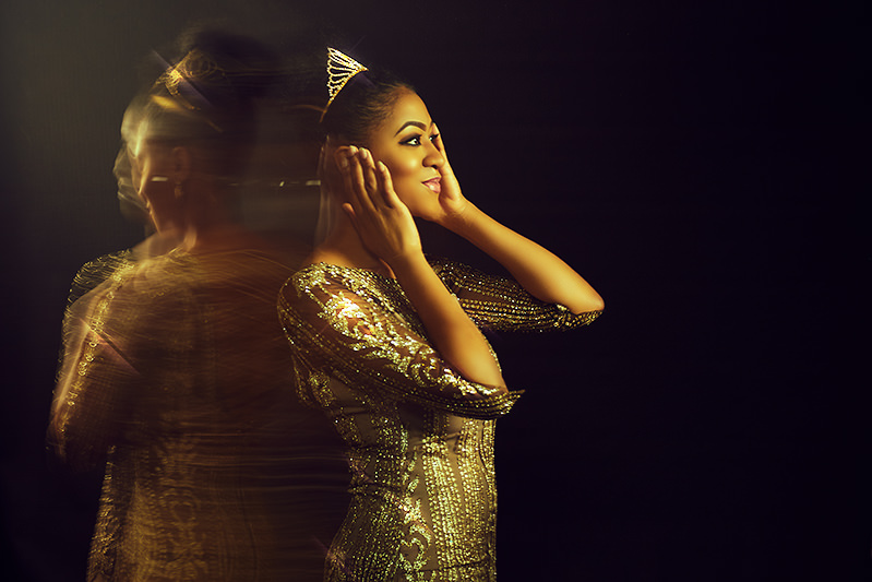 Woman in a tiara and gold dress with hands at her head, smiling, a blurred double trailing behind her