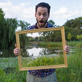 Man holding an empty ornate gold picture frame up to the camera in front of a pond and trees, example for creating a see-through frame effect in Photoshop