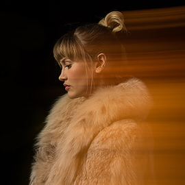 Woman with blonde hair in a fur coat photographed in profile with motion blur and warm lighting, thumbnail for the Photo Editing 101 course