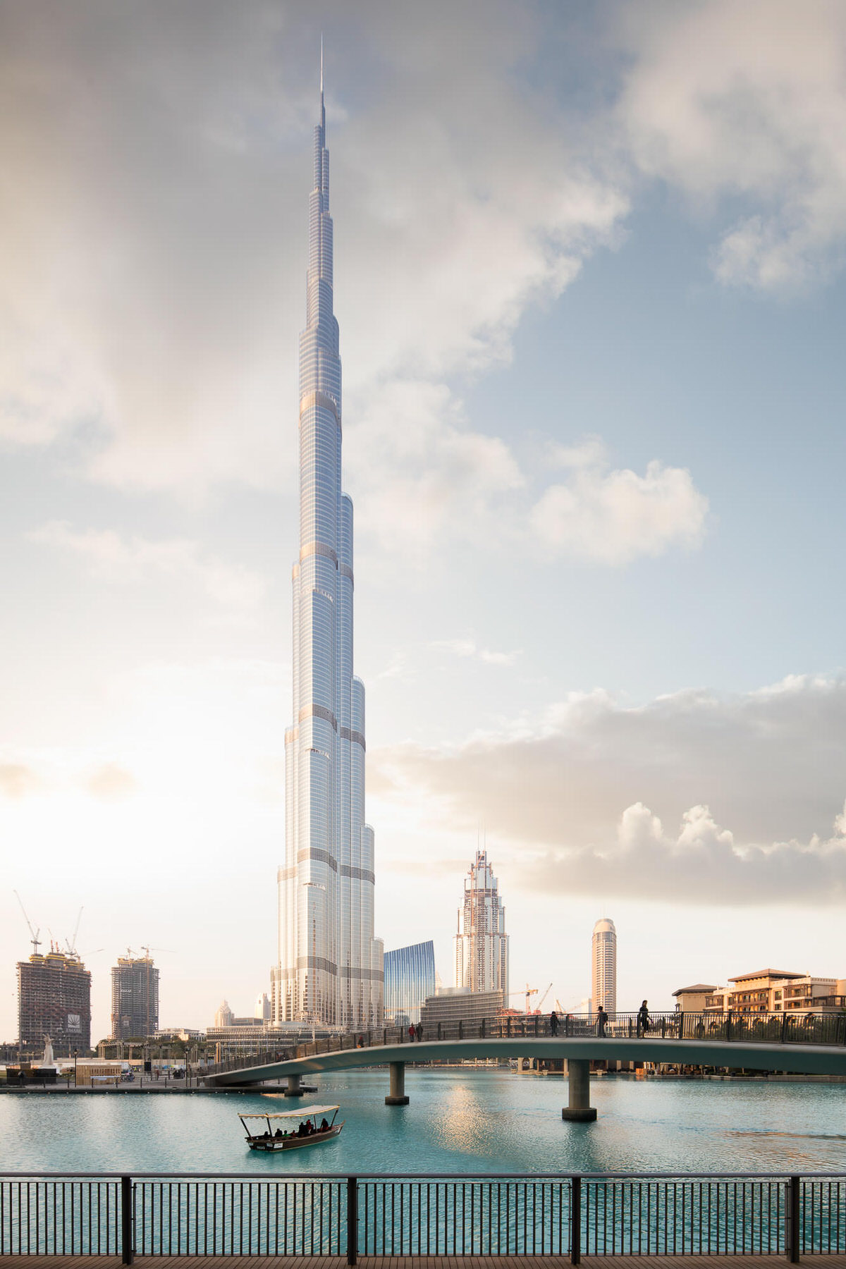 Burj Khalifa rising above a turquoise lake with a footbridge and a small boat in soft evening light