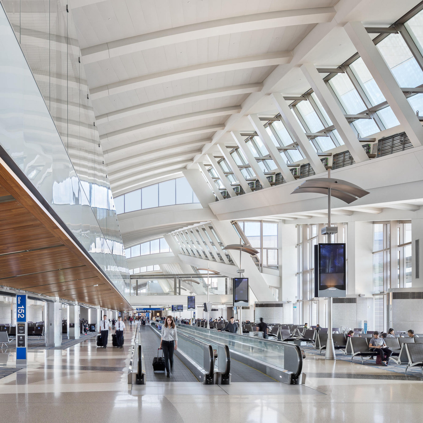 Bright airport terminal concourse with a curved white roof, skylights and moving walkways, a traveler pulling a suitcase