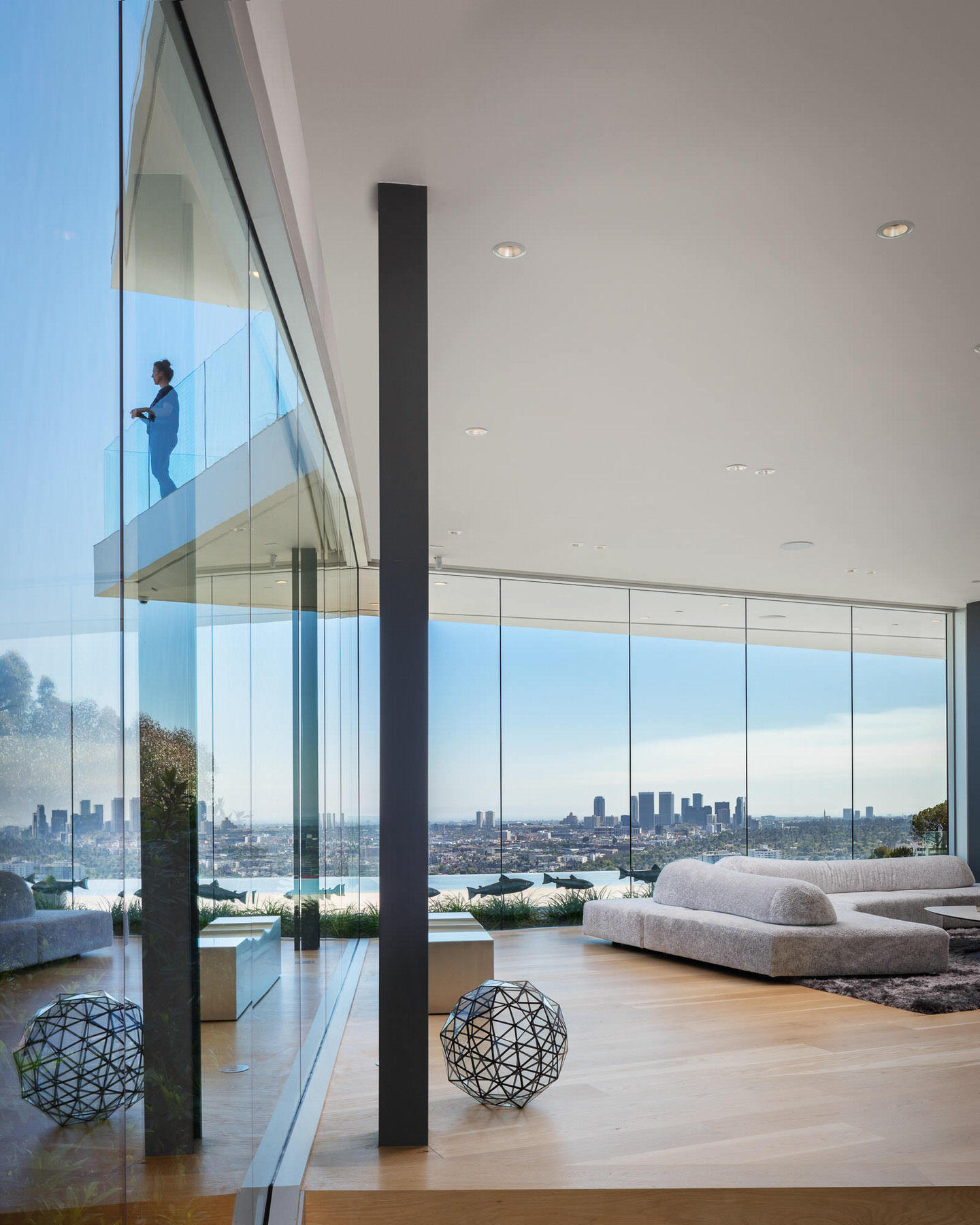 Glass-walled hillside home with a white sofa and city views, a person standing on the balcony above