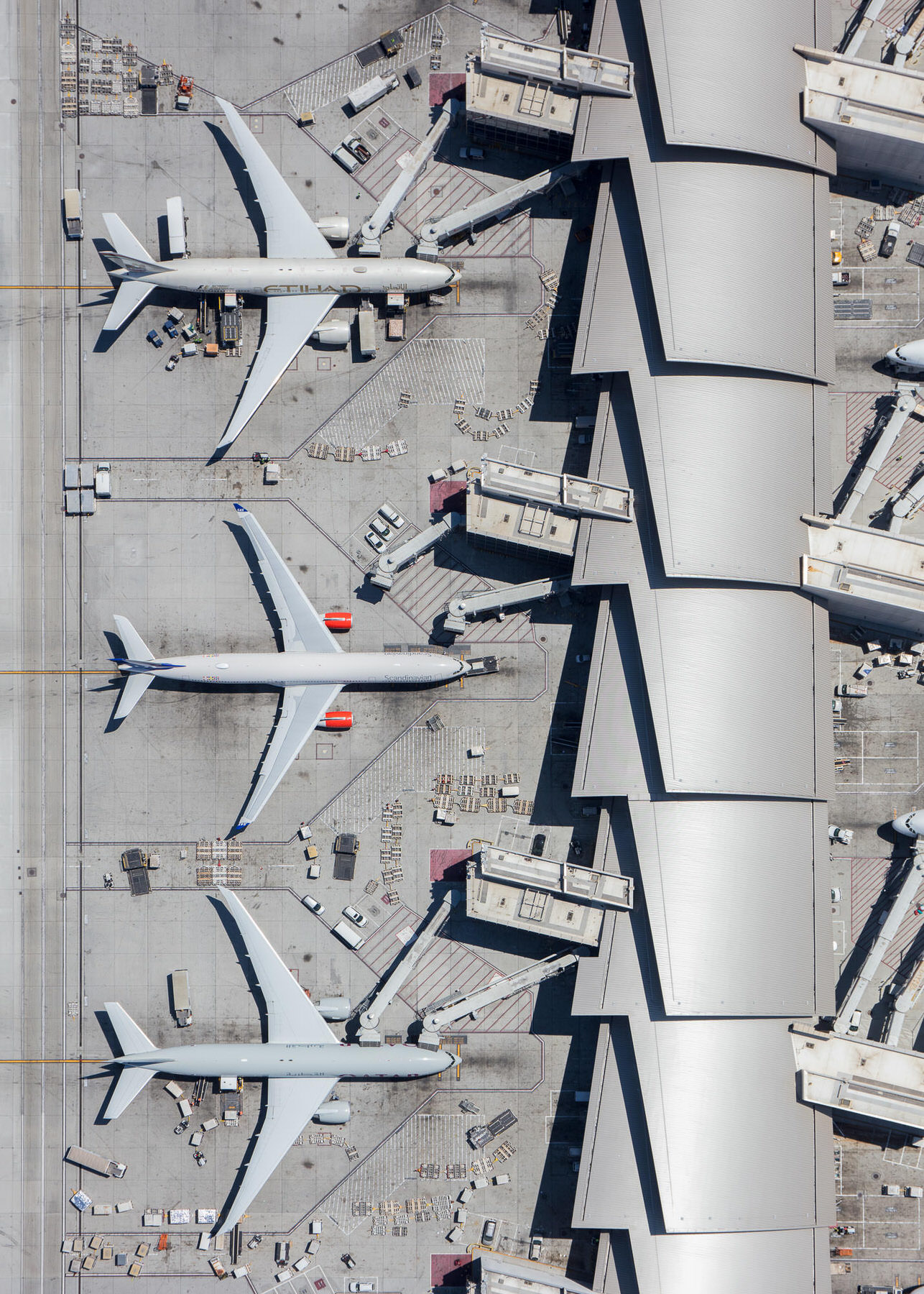 Aerial view straight down on three white jets parked at terminal gates with jet bridges and ground vehicles