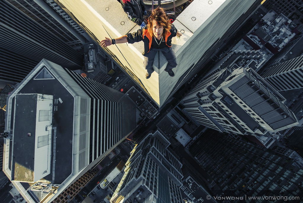 Ben Von Wong in an orange vest sitting on the edge of a skyscraper roof, shot from above with towers and streets far below