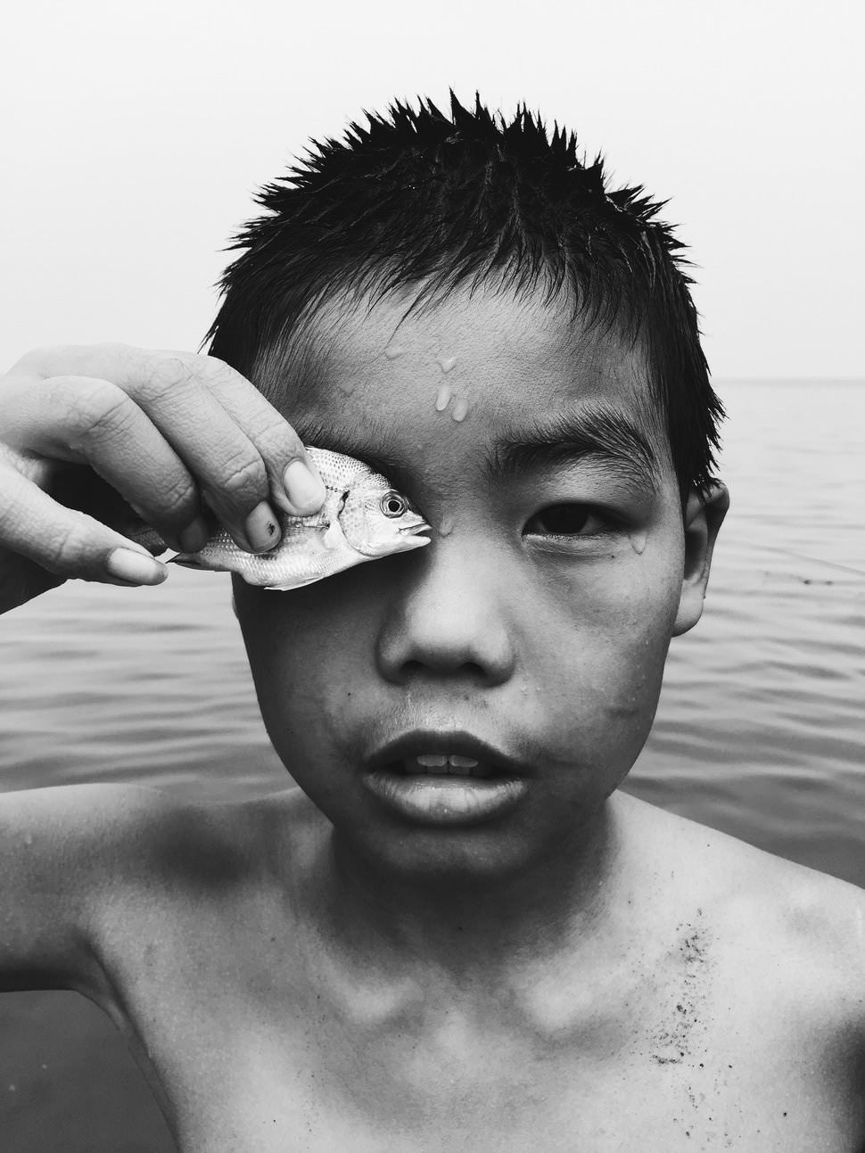 Eye to Eye: black-and-white portrait of a wet-haired boy by the sea holding a small fish over one eye