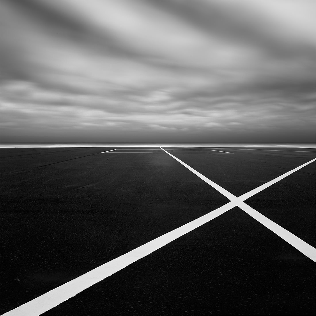 .Rhythm: black-and-white photo of white lines crossing on dark tarmac under streaked clouds