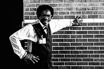 Black-and-white portrait of a smiling museum guide in a vest and glasses leaning on a brick wall
