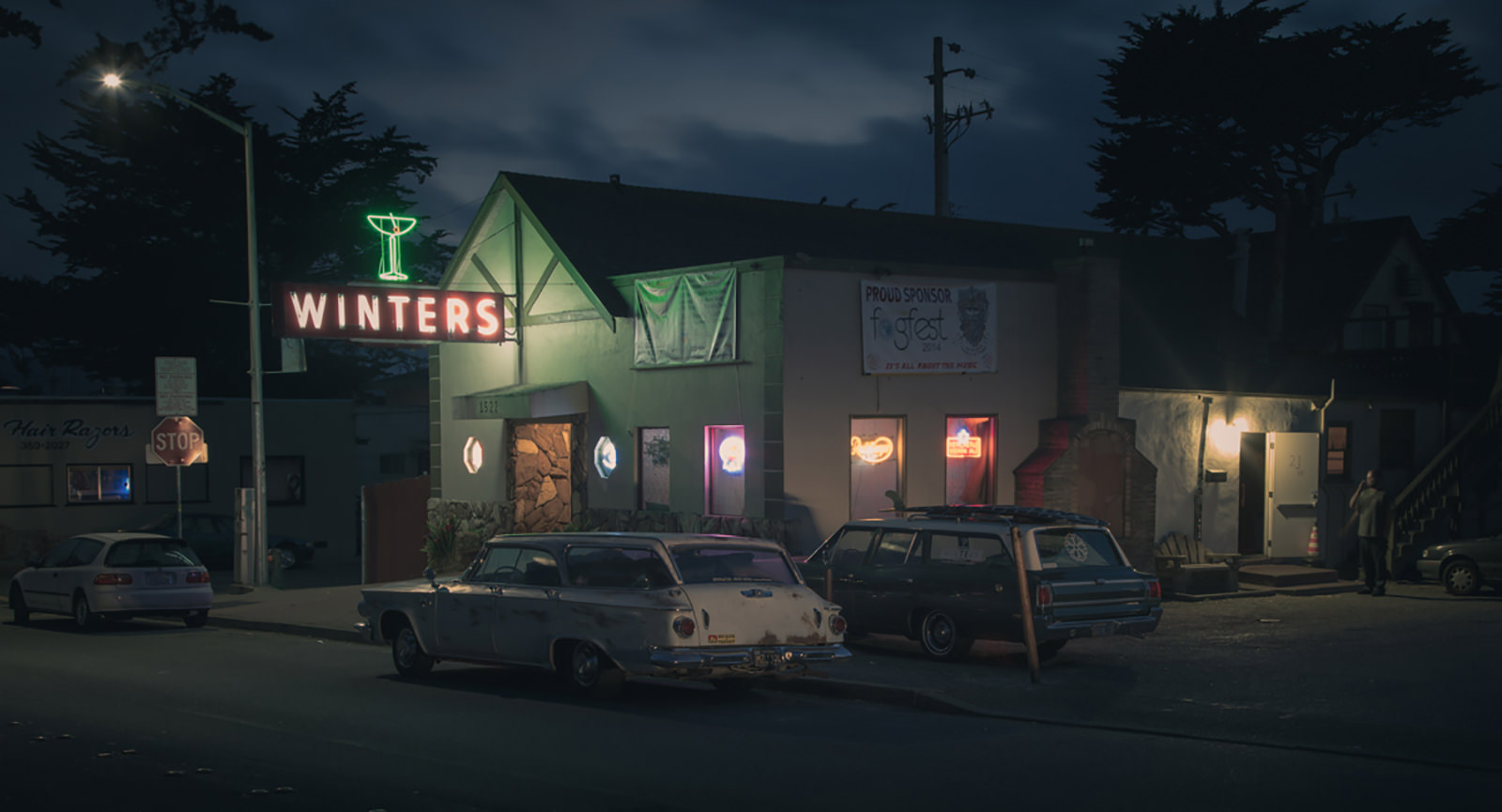 Wider night view of the Winters bar with its neon sign, two vintage station wagons parked out front and a man by a side door