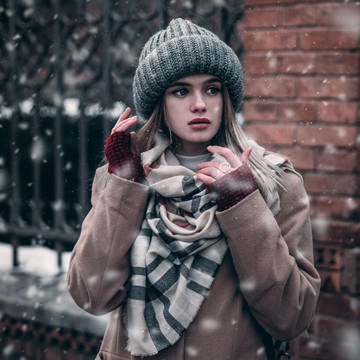 Woman in a knit beanie and plaid scarf standing by a brick wall and iron gate in falling snow, example for creating a custom brush in Photoshop