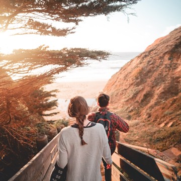 Couple walking down a wooden boardwalk toward a sunlit beach between coastal cliffs and pine trees, example photo for editing faster with custom keyboard shortcuts in Photoshop