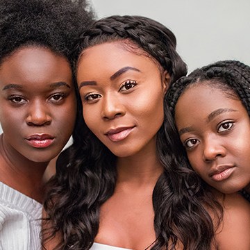 Three women with dark hair styled differently — afro, braided crown, and long waves — posed close together for a sharpness comparison between Photoshop and Lightroom