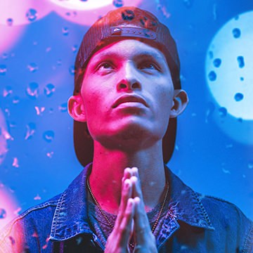 Man in a backwards cap and denim jacket with hands clasped in prayer, lit with pink and blue tones and a bokeh water-droplet texture overlay