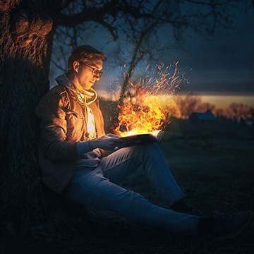 Man sitting under a tree at dusk with a glowing fire coming from an open book, demonstrating how to create realistic fire in Photoshop