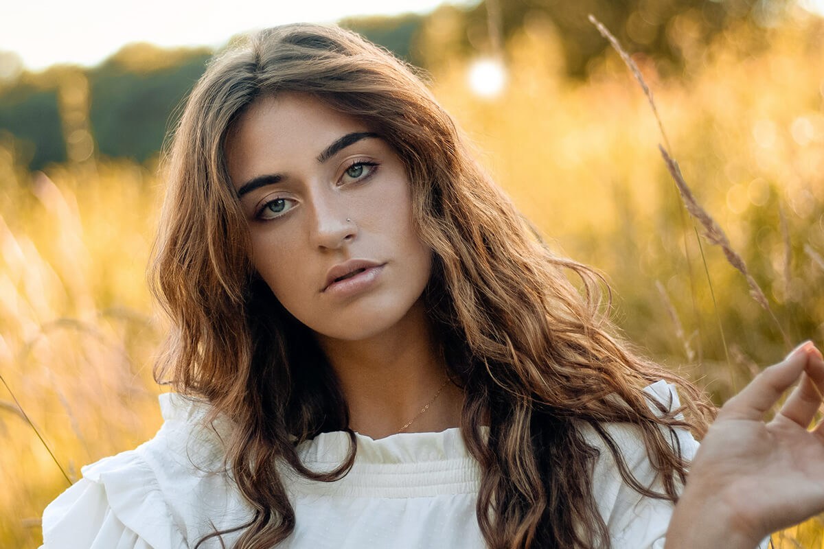 Total image makeover in Photoshop, part 2: a woman with long wavy brown hair poses in a golden-lit field wearing a white top after object removal, sky replacement and color grading.