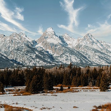 Snow-capped mountain range with wispy clouds above a pine forest, thumbnail for a tutorial on adding realistic clouds with Photoshop's Blend If