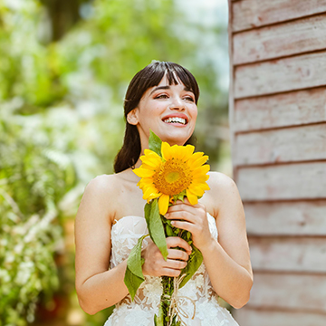 Smiling woman with bangs holding a sunflower near a wooden building, thumbnail for a tutorial on using Lens Blur in Photoshop