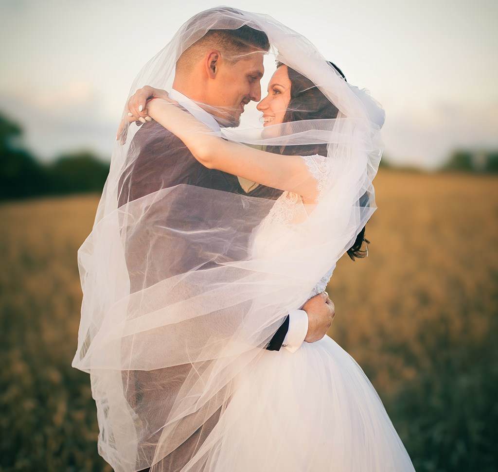 The same bride and groom embracing under a veil in a wheat field after warm, dreamlike wedding-style color grading