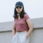 A woman in a cap and sunglasses by a white wall