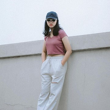 A woman in a pink crop top, white pants and a cap leaning against a wall in a casual streetwear portrait — How to Use the Clone Stamp Tool in Photoshop tutorial thumbnail