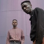 Two men in earth-toned clothes against a curved metal wall