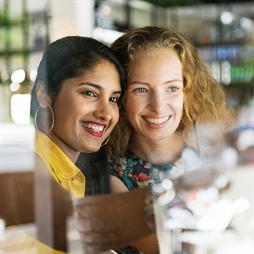 Two women smiling and looking through a reflective café window — How to Remove Reflections From Faces in Photoshop 2026 tutorial thumbnail
