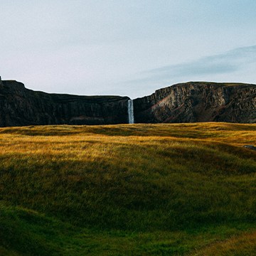 A dramatic grassy cliff-top landscape with a waterfall in the distance under golden evening light — Fundamentals of Composition & Light in Photoshop tutorial thumbnail