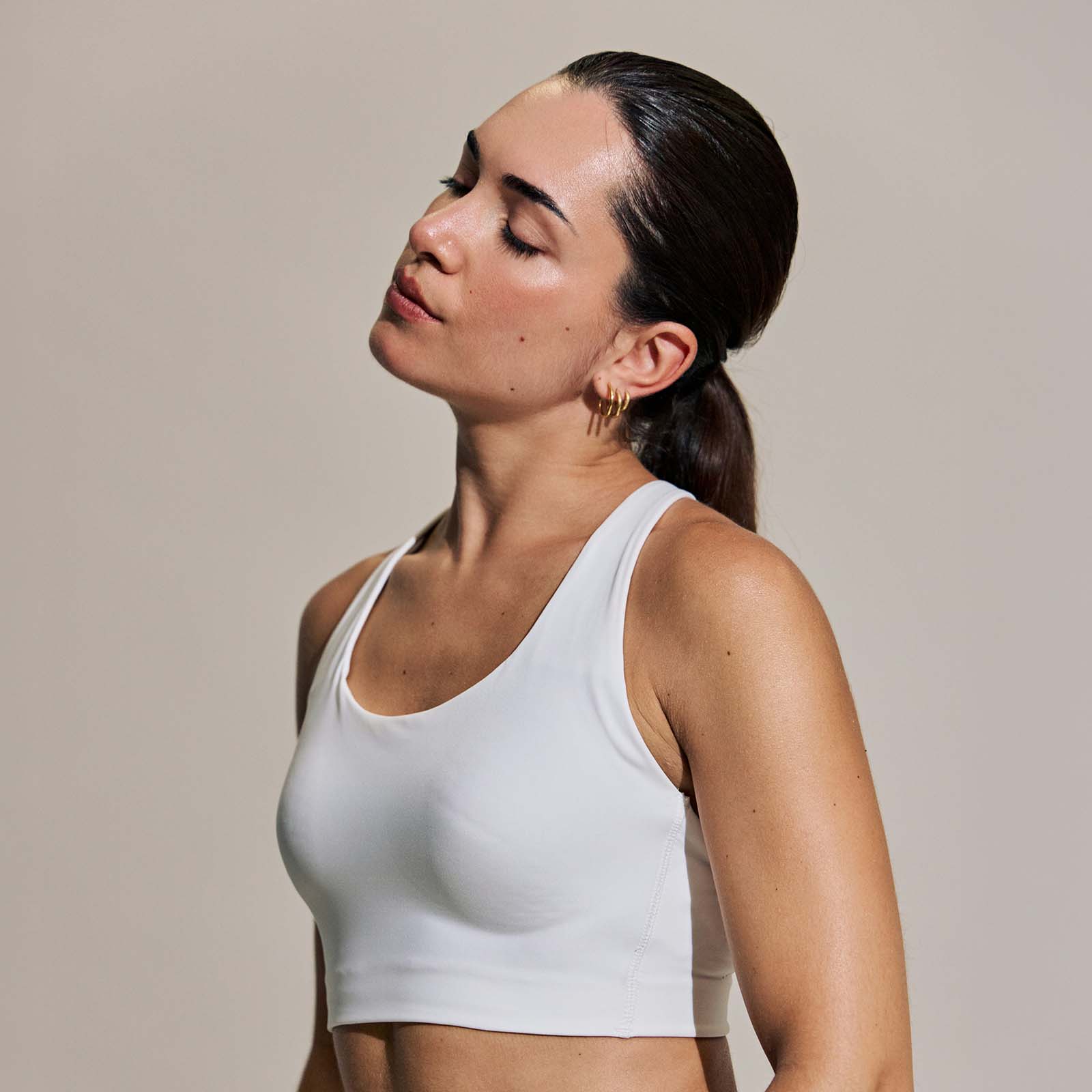 A profile portrait of a woman in a white tank top with her eyes closed and hair pulled into a low ponytail, showing visible skin blemishes and moles before any retouching