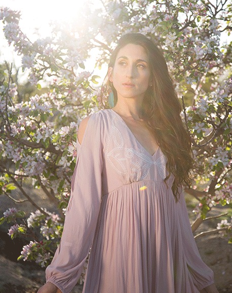 Woman in a pale pink gown standing among sunlit foliage