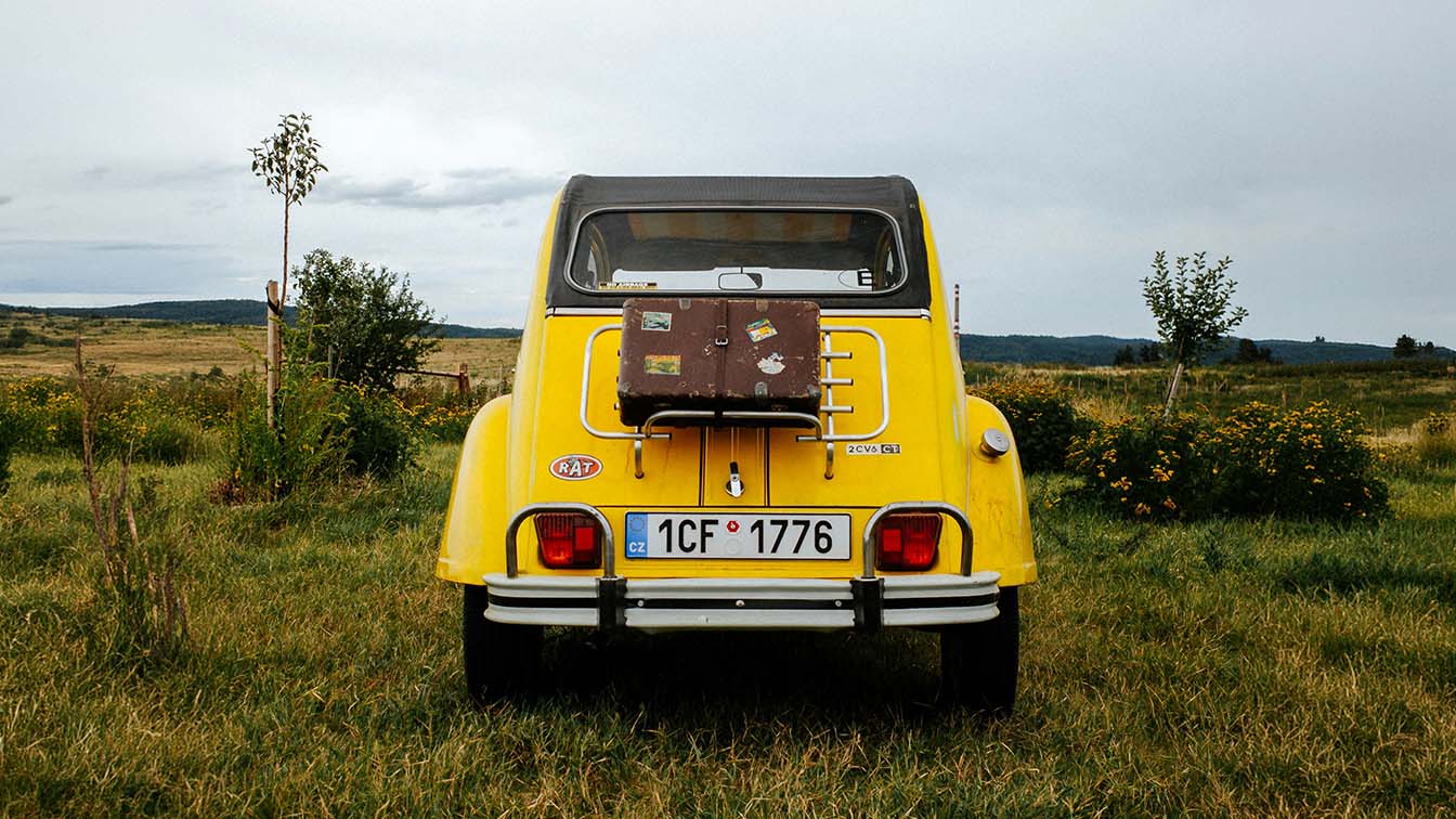 The back of a yellow vintage car with a suitcase strapped to it, parked in a grassy field